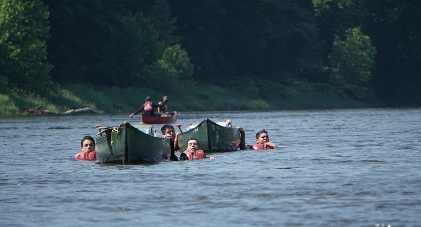 A group of people wearing life jackets float in water alongside empty canoes 